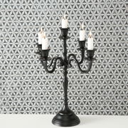 Schwarzer Kerzenleuchter Victoria mit fünf Armen und brennenden Kerzen, frontal vor gemustertem Hintergrund.