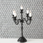 Schwarzer Kerzenleuchter Victoria mit fünf Armen und brennenden Kerzen, frontal vor gemustertem Hintergrund.