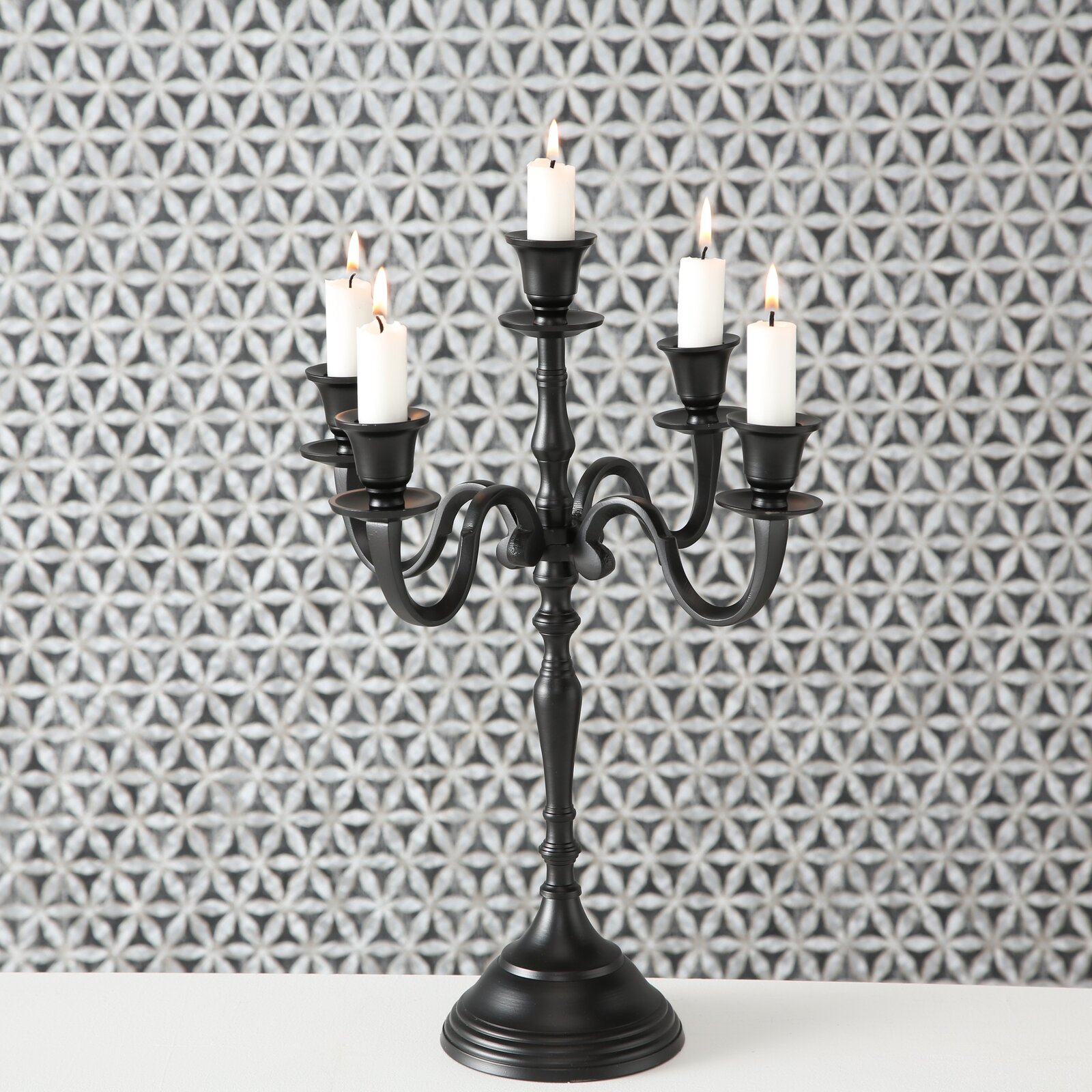 Schwarzer Kerzenleuchter Victoria mit fünf Armen und brennenden Kerzen, frontal vor gemustertem Hintergrund.