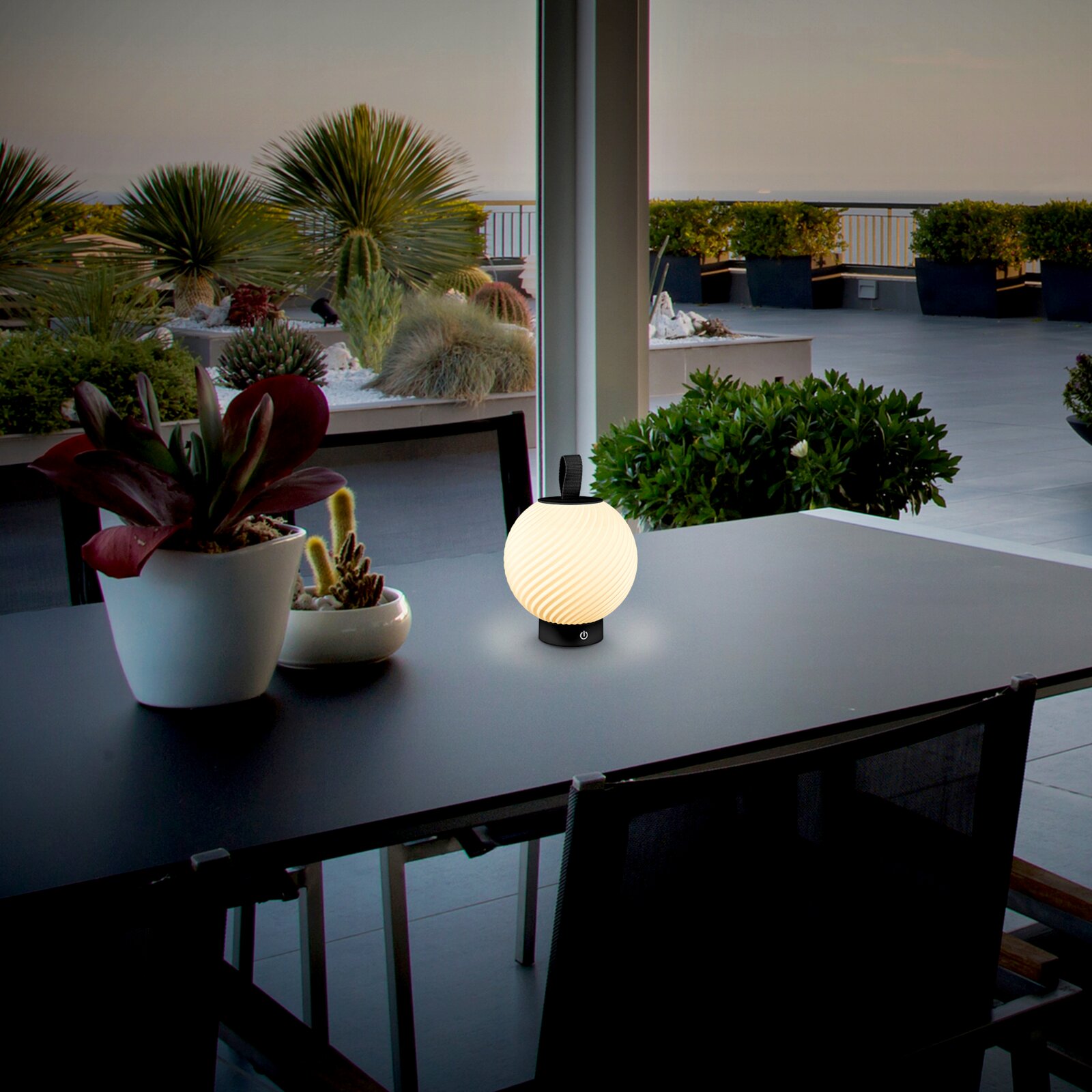 Briloner Akku-Tischleuchte BRILO UNPLUGGED Leuchtende, kugelförmige LED-Tischlampe mit geriffeltem Diffusor und schwarzem Sockel auf dunklem Outdoor-Tisch auf einer Dachterrasse mit Pflanzen und Meerblick, aufgenommen aus frontaler Tischhöhe in leichter Seitenperspektive.