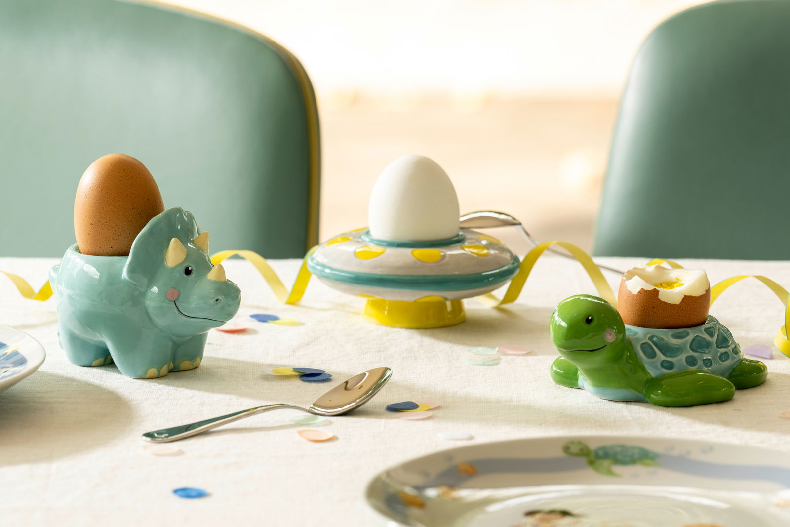 Drei bunte Eierbecher in Form eines Dinosauriers, einer Schildkröte und eines UFOs auf einem gedeckten Tisch, aus der Perspektive von schräg oben fotografiert.