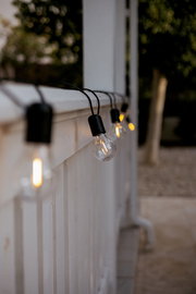 Leuchtende Outdoor-Lichterkette mit klaren Glühbirnen an einem weißen Geländer, seitliche Nahaufnahme aus schräger Perspektive entlang des Geländers