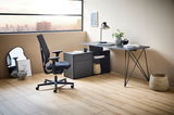 Moderner Winkelschreibtisch mit Kommode in einem stilvollen Büro, seitliche Perspektive. Der Schreibtisch hat eine schwarze Oberfläche und filigrane Metallbeine. Eine Kommode mit Schubladen und offenen Fächern bietet zusätzlichen Stauraum. Ein ergonomisch