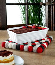 Quadratischer Untersetzer LANA ROSSO aus Filzkugeln in Rot, Weiß und Schwarz, auf einem Holztisch mit einer rechteckigen weißen Auflaufform und einem Stück Kuchen im Vordergrund, seitliche Perspektive.