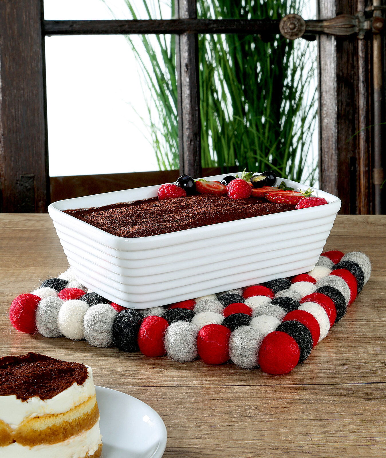 Quadratischer Untersetzer LANA ROSSO aus Filzkugeln in Rot, Weiß und Schwarz, auf einem Holztisch mit einer rechteckigen weißen Auflaufform und einem Stück Kuchen im Vordergrund, seitliche Perspektive.