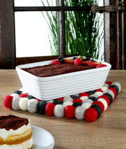 Quadratischer Untersetzer LANA ROSSO aus Filzkugeln in Rot, Weiß und Schwarz, auf einem Holztisch mit einer rechteckigen weißen Auflaufform und einem Stück Kuchen im Vordergrund, seitliche Perspektive.