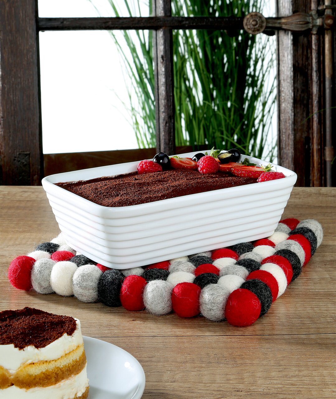 Quadratischer Untersetzer LANA ROSSO aus Filzkugeln in Rot, Weiß und Schwarz, auf einem Holztisch mit einer rechteckigen weißen Auflaufform und einem Stück Kuchen im Vordergrund, seitliche Perspektive.