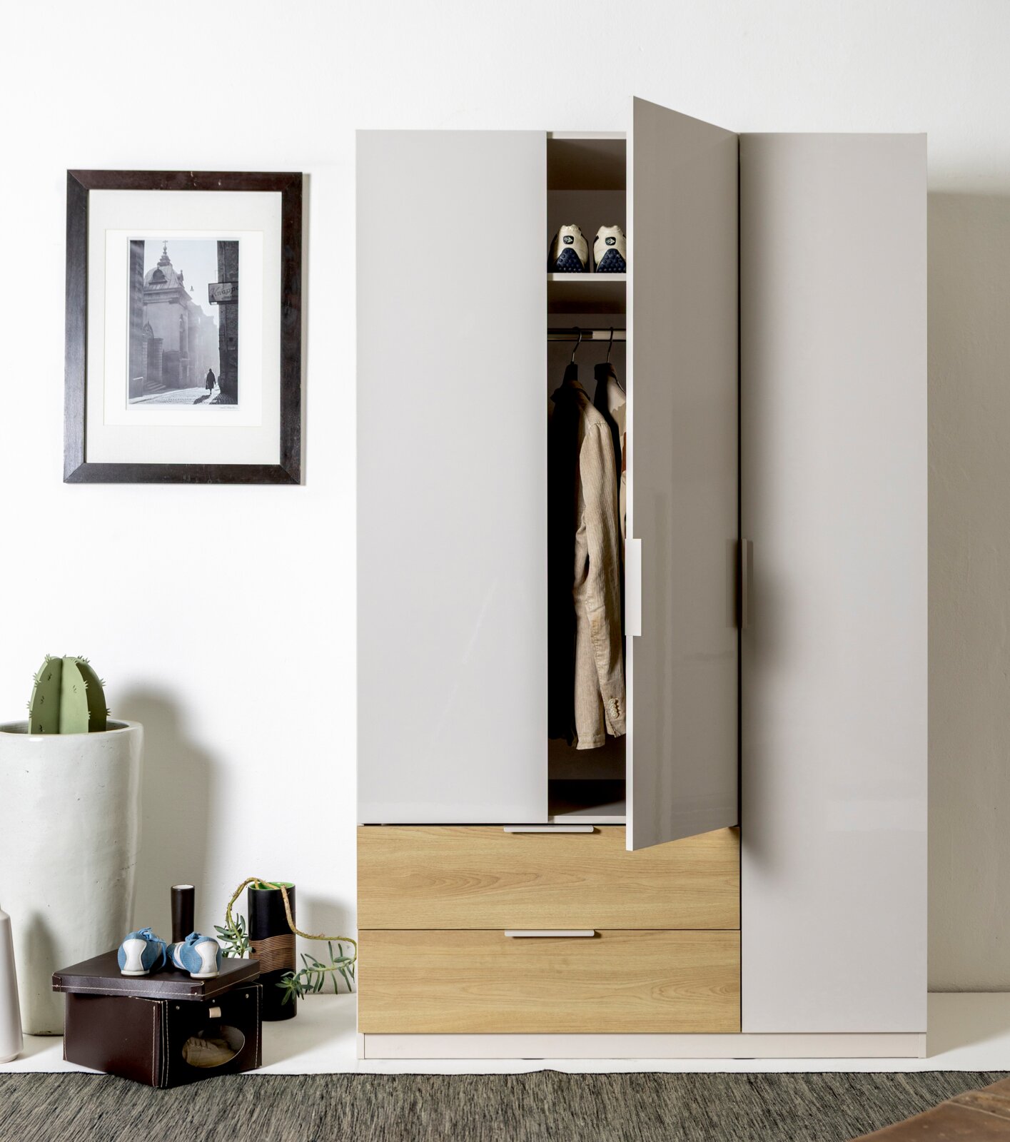 Moderne kledingkast in wit met houten lades, één deur open met zichtbare kledingstang en opbergvak, vooraanzicht in licht schuin perspectief