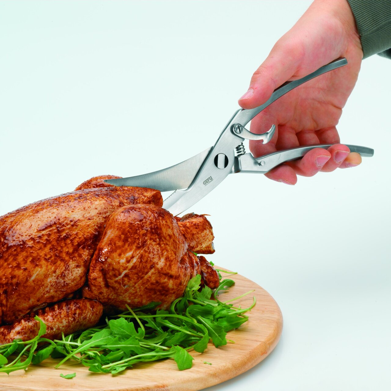 Geflügelschere mit Feder in der Hand, die ein gebratenes Hähnchen auf einem Holzbrett schneidet, seitliche Perspektive