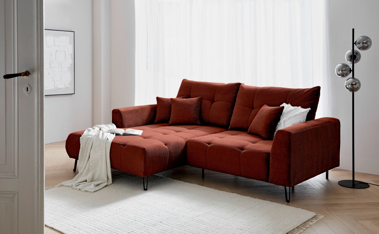 Rode hoekbank met poef aan de linkerkant, versierd met kussens en een deken, in een moderne woonkamer met een lichte houten vloer en een staande lamp, gezien vanaf links vooraan.