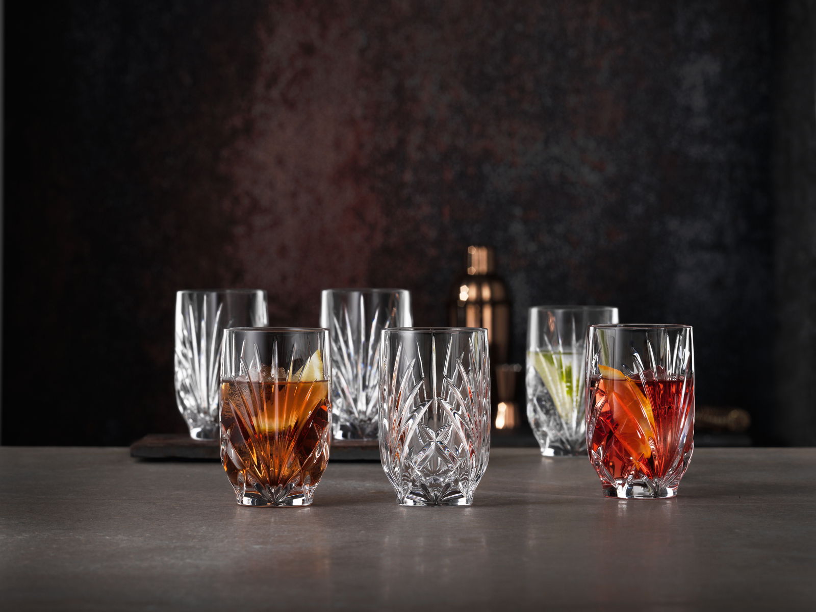 Nachtmann Saftgläser PALAIS Sechs elegante Kristallgläser mit facettiertem Design, gefüllt mit bunten Getränken, auf einer dunklen Oberfläche vor einem dunklen Hintergrund, aus einer leicht erhöhten Perspektive fotografiert.