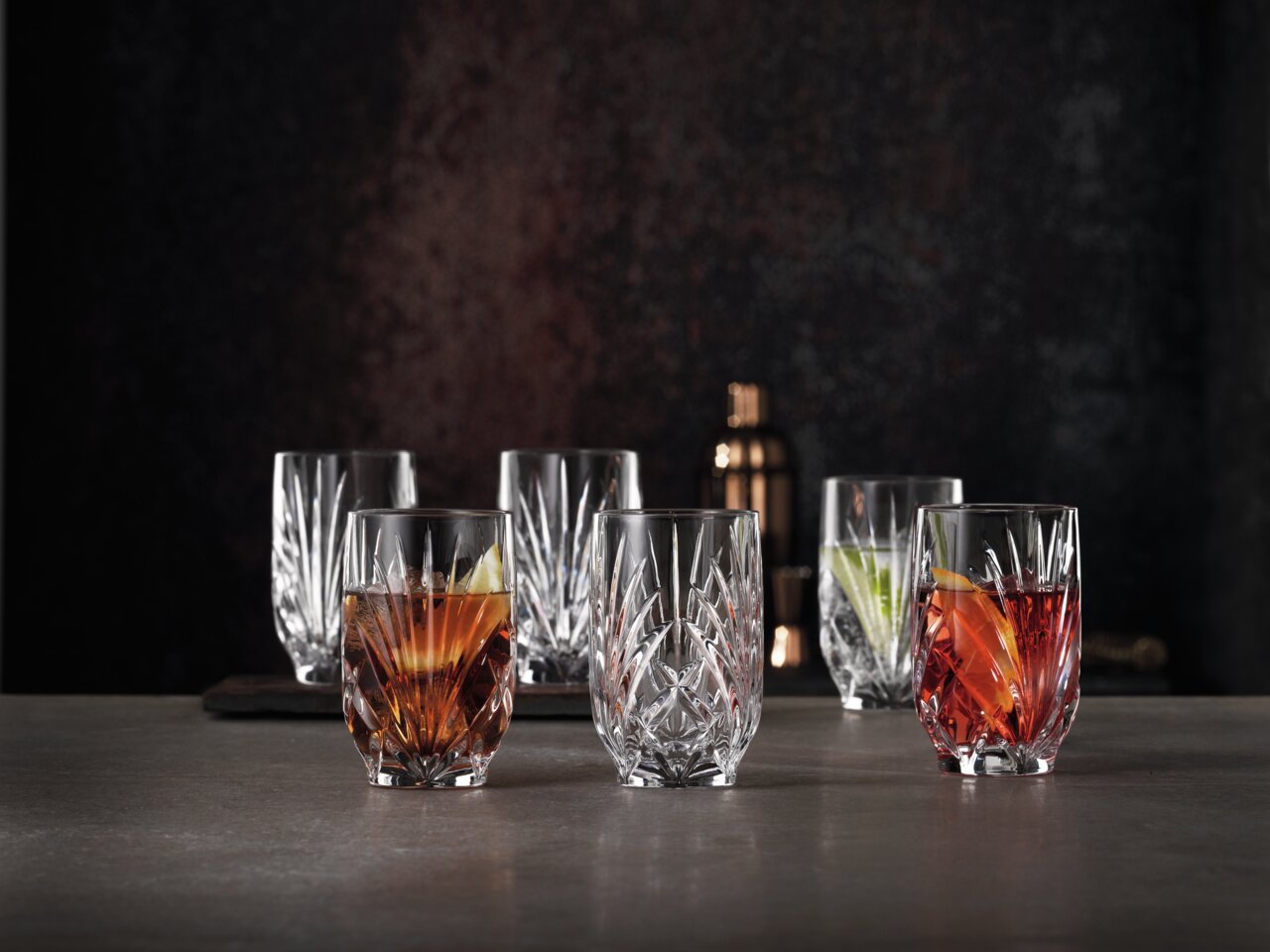 Nachtmann Saftgläser PALAIS Sechs elegante Kristallgläser mit facettiertem Design, gefüllt mit bunten Getränken, auf einer dunklen Oberfläche vor einem dunklen Hintergrund, aus einer leicht erhöhten Perspektive fotografiert.