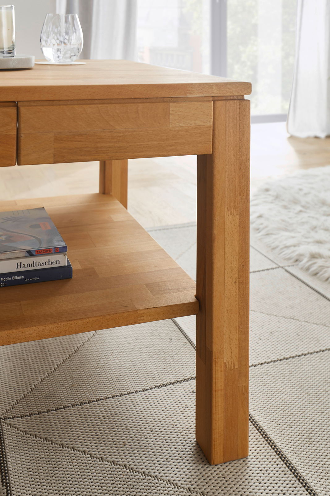 Nahaufnahme eines modernen Couchtisches aus Holz mit klaren Linien und einer unteren Ablage, auf der Bücher liegen. Der Tisch steht auf einem hellen Teppich in einem gut beleuchteten Raum.