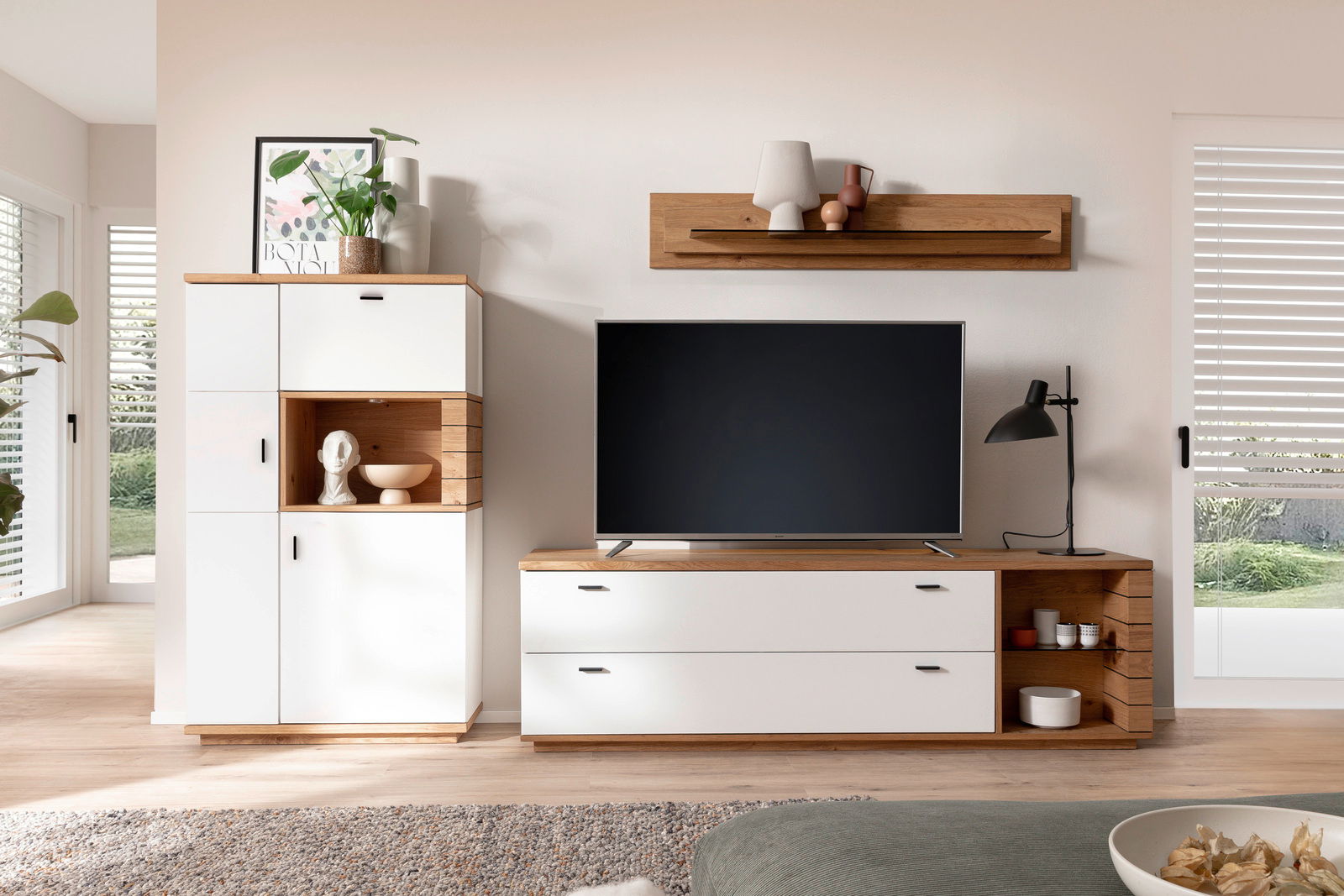 Novel Wandkombination MANITOBA Moderne Wandkombination in einem Wohnzimmer mit weißem und holzfarbenem Design, bestehend aus einem hohen Schrank, einem TV-Schrank und einem Wandregal. Perspektive von vorne.