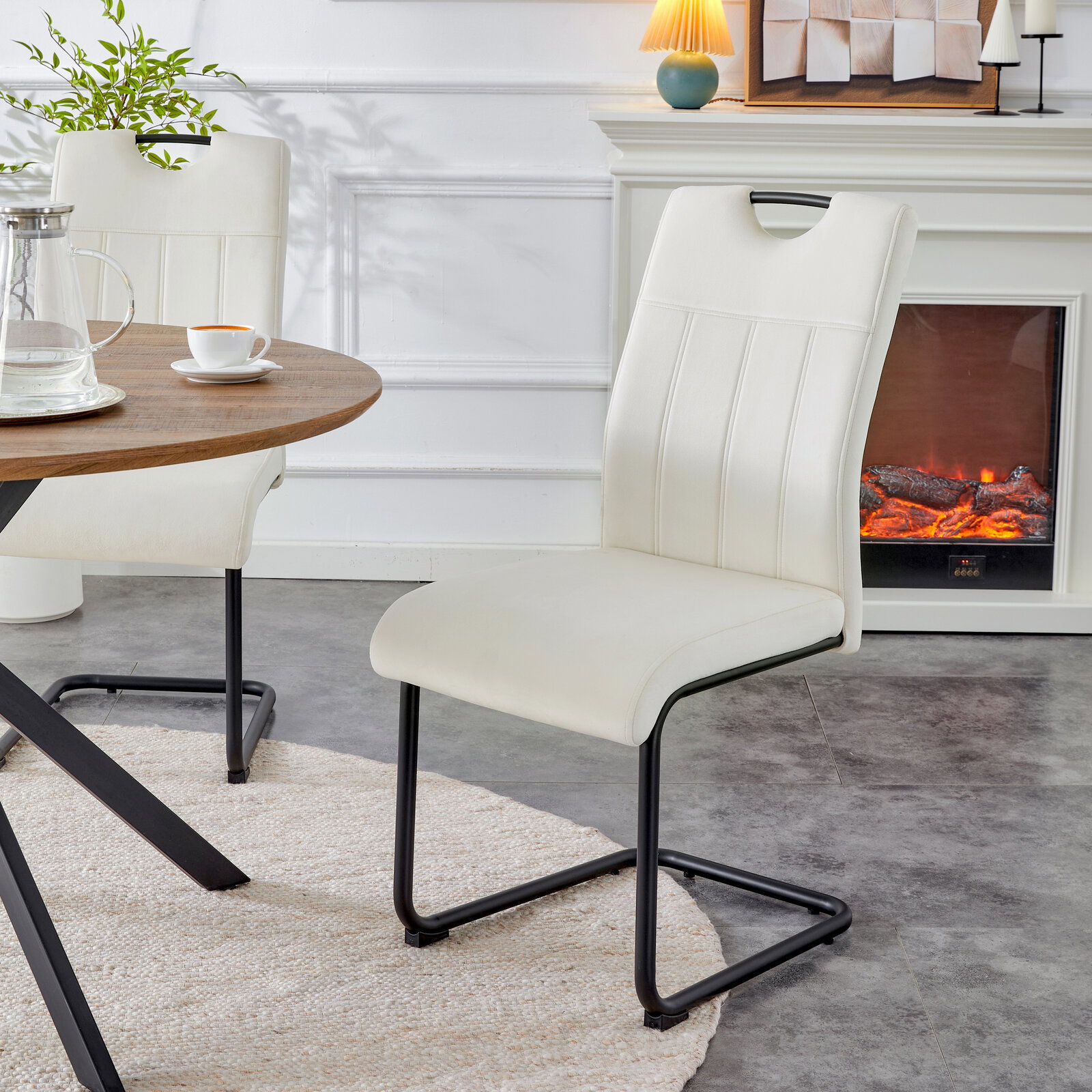 Cremeweißer Polster-Esszimmerstuhl mit Steppnähten und schwarzem Metall-Schwinggestell, seitliche Frontansicht im Wohn-/Essbereich neben rundem Holztisch und Kamin