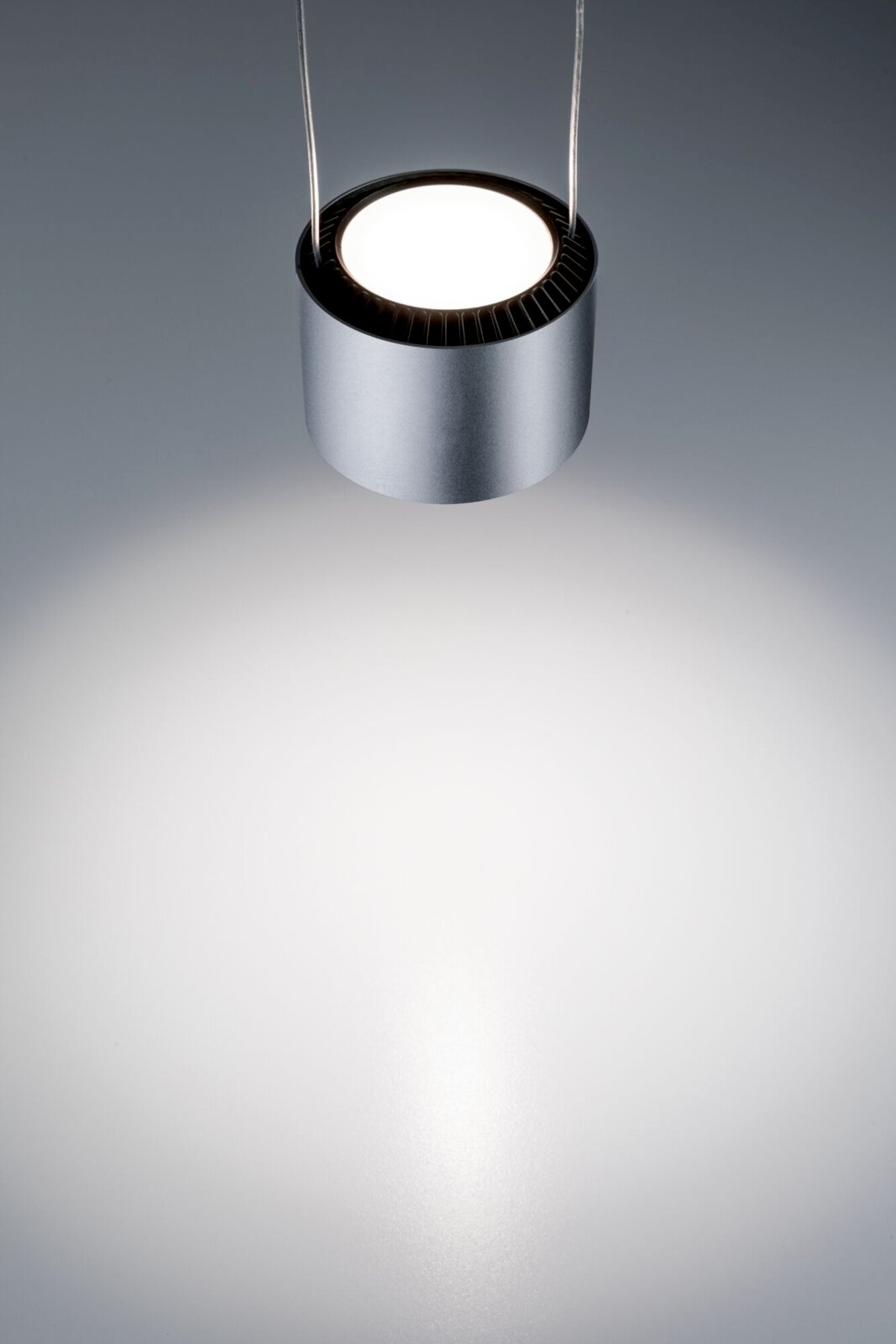 Moderne, zylindrische Hängeleuchte in silberner Farbe, von oben fotografiert, mit hellem Lichtstrahl auf einer grauen Oberfläche.