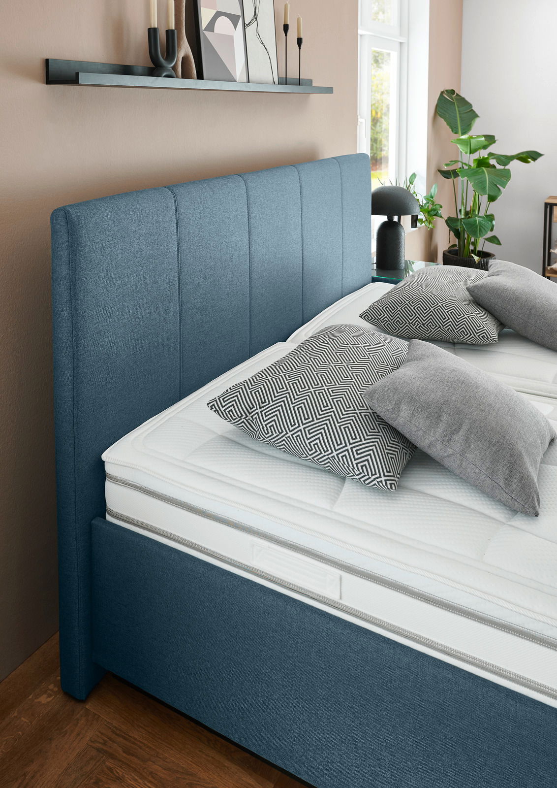 Seitliche Perspektive auf ein modernes Polsterbett mit blauem Kopfteil, dekoriert mit gemusterten Kissen und umgeben von stilvollen Accessoires.