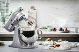 Küchenmaschine mit Edelstahlschüssel, seitliche Perspektive, in einer modernen Küche mit Zutaten im Hintergrund.