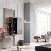 Modernes Wohnzimmer mit schlanker, weißer Bogen-Stehlampe neben Sessel und Vitrine, Innenaufnahme in frontaler Seitenansicht.