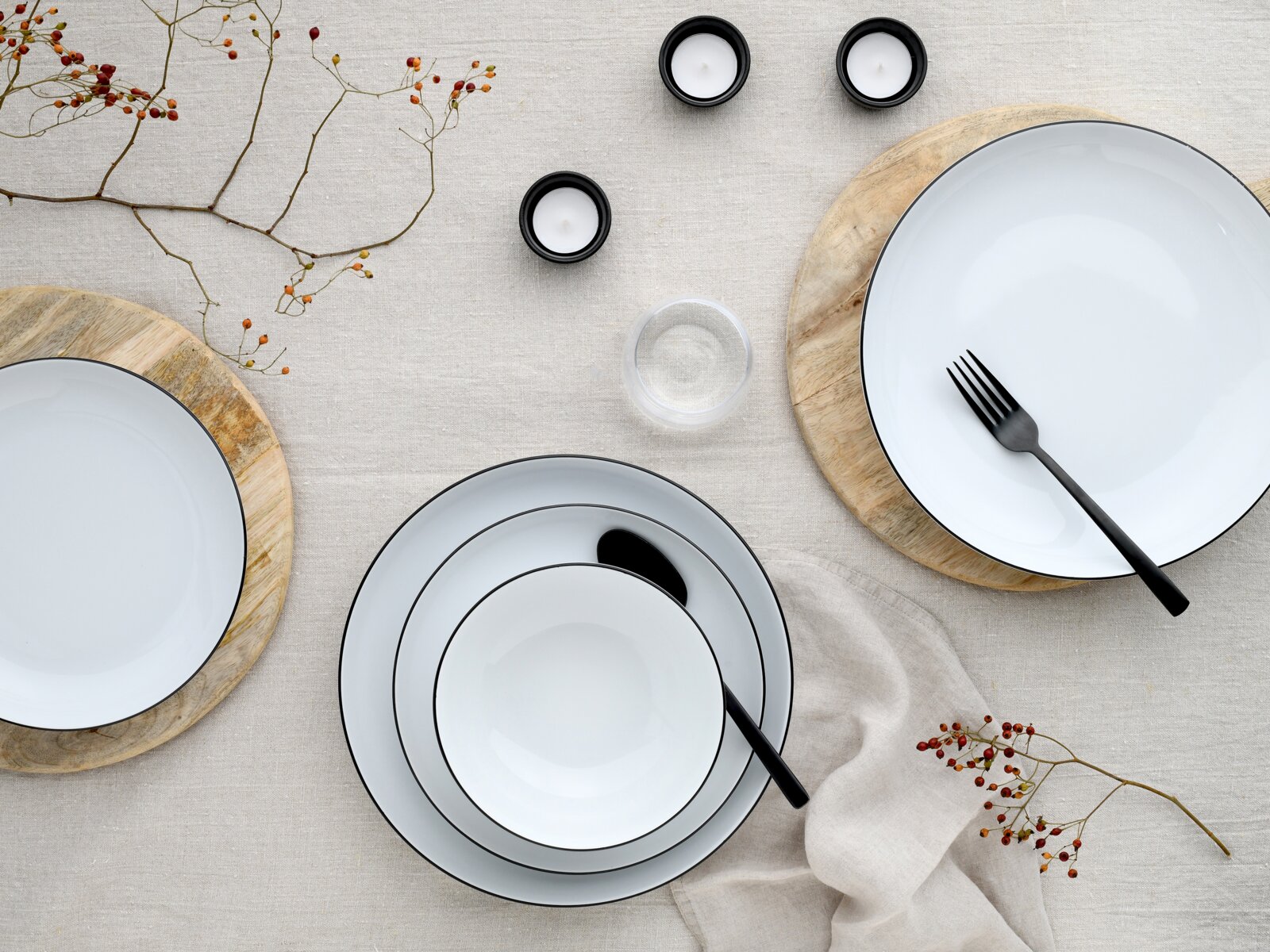 Draufsicht auf ein minimalistisch gedecktes Tischarrangement mit weißen Tellern und Schalen mit schwarzem Rand, schwarzem Besteck, Teelichtern, Glas, Holzuntersetzern und Zweigen mit roten Beeren auf hellem Leinentuch.