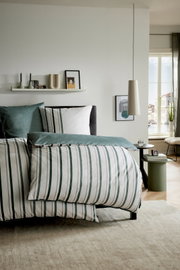 Gestreept beddengoed in wit met groenblauwe strepen op een opgemaakt bed in een lichte slaapkamer, gefotografeerd vanuit een schuine vooraanzicht-perspectief.