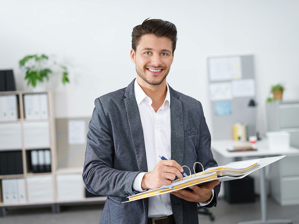Ein lächelnder Mann im Büro hält einen Ordner und schreibt mit einem Stift.