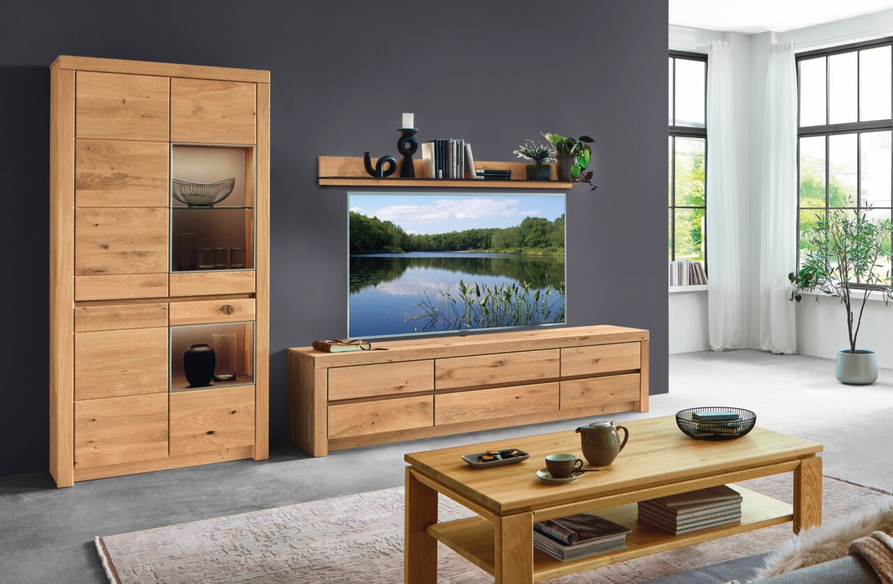 Wöstmann Markenmöbel Wandcombinatie SOLEO 3000 Uitzicht op de woonkamer met de moderne SOLEO 3000-wandcombinatie van het merk Wöstmann in licht hout, bestaande uit een vitrinekast, een tv-meubel en een wandrek, met decoratieve elementen en uitzicht op grote ramen.