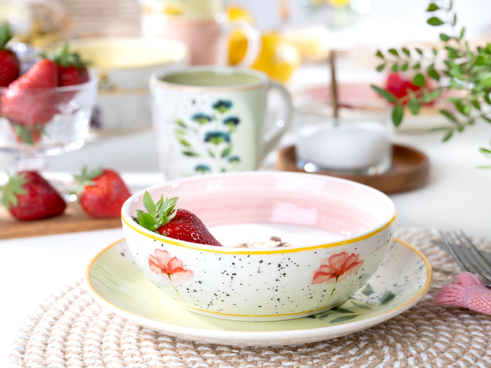 Leicht von oben fotografierte Keramikschale mit rosa Innenfläche, floralem Dekor und gelbem Rand auf passendem Teller, gefüllt mit Joghurt, Müsli und einer Erdbeere.