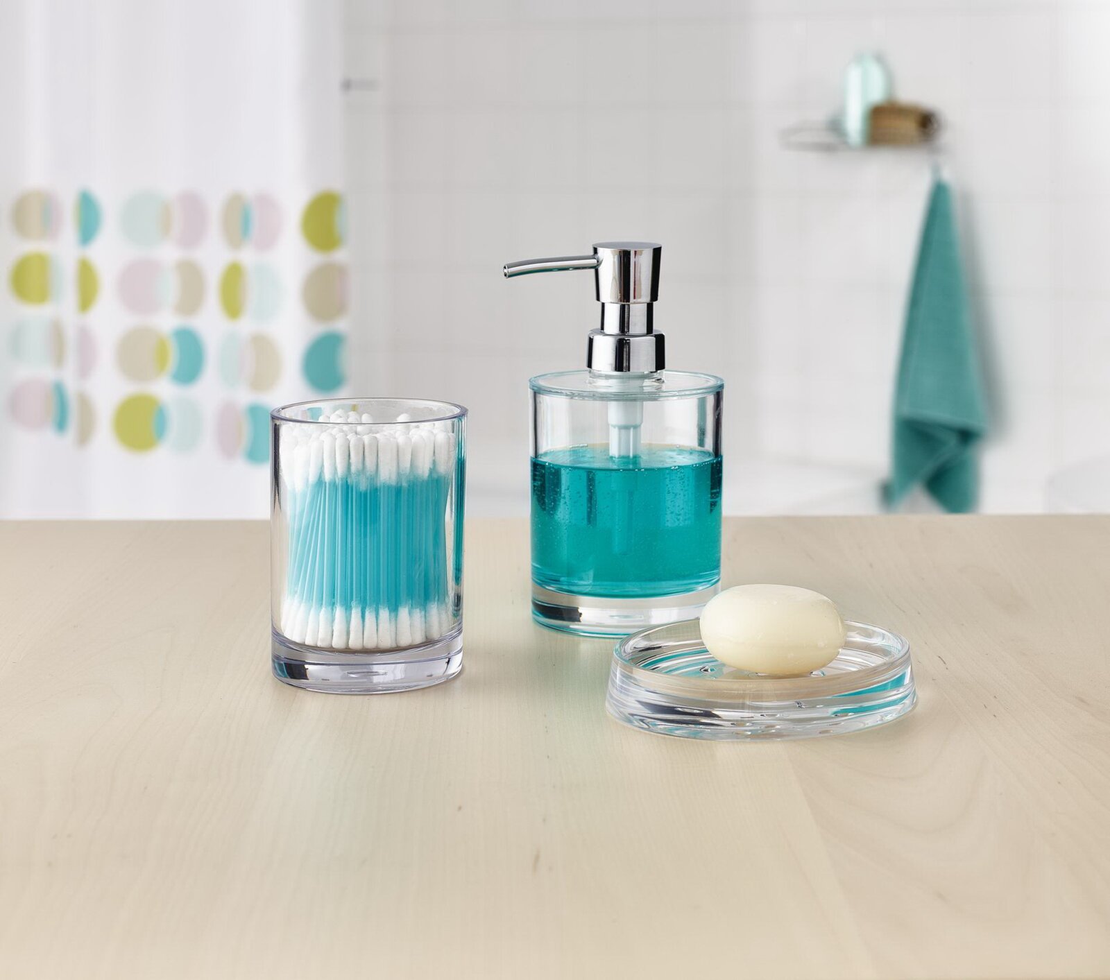 Transparentes Bad-Accessoire-Set aus Glas: Seifenspender mit türkisfarbener Flüssigseife, Wattestäbchen-Behälter und Seifenschale mit Seifenstück, frontal aus leicht erhöhter Perspektive auf einem hellen Waschtisch.