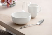 Weiße Müslischale Afina 550ml auf einem Tisch, seitliche Perspektive, neben einer Tasse und einem Löffel.