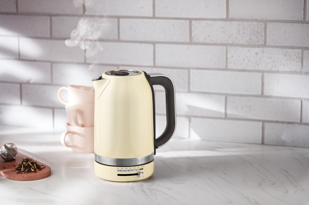 KitchenAid Wasserkocher Beiger Wasserkocher mit schwarzem Griff, seitlich auf einer Küchenarbeitsplatte vor einer weißen Ziegelwand platziert, mit Dampf, der aus dem Ausguss aufsteigt.
