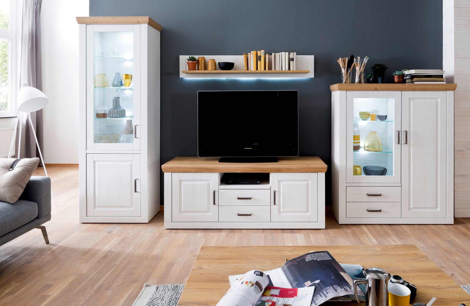 Wandmeubel in landhuisstijl in wit met eikenlook: links een hoge vitrinekast met glazen deur en verlichting, in het midden een tv-lowboard met televisie en wandplank, rechts een dressoir met vitrinekast en lades; vooraanzicht in de woonkamer