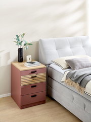 Rollcontainer in warmem Rotton mit drei Schubladen, Holzoberfläche und schwarzen Griffen, seitliche Perspektive neben einem Bett.