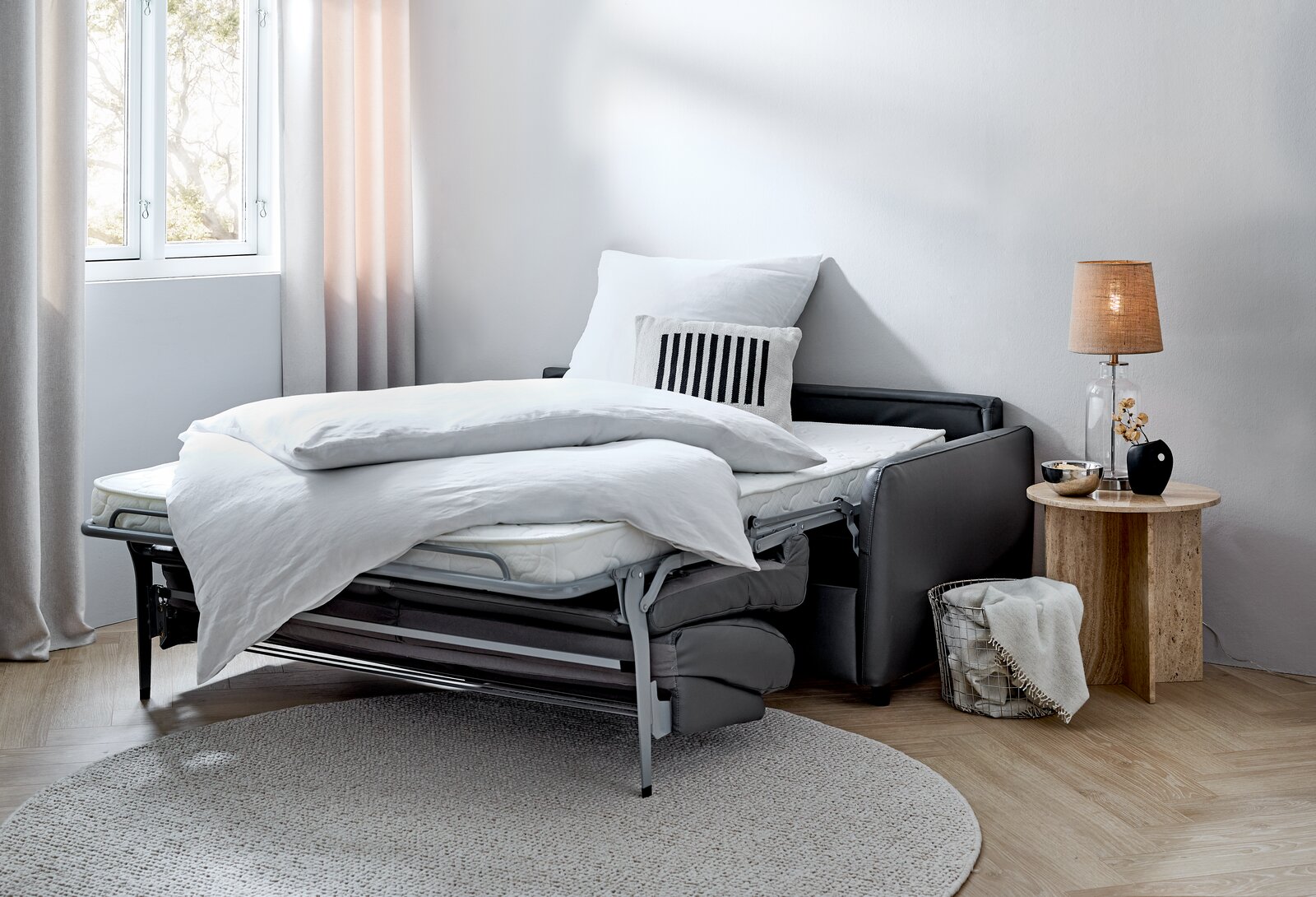 Funktionssofa in ausgeklappter Bettposition, seitliche Perspektive, mit weißer Bettwäsche und Kissen, neben einem runden Beistelltisch mit Lampe.