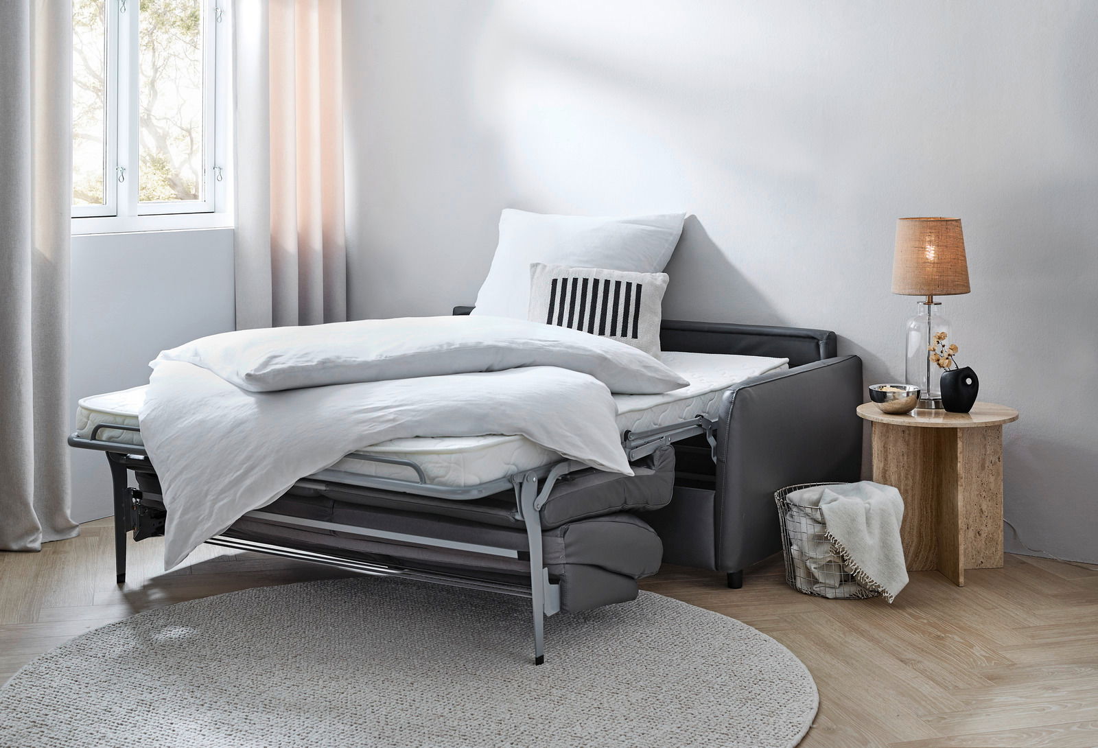 Funktionssofa in ausgeklappter Bettposition, seitliche Perspektive, mit weißer Bettwäsche und Kissen, neben einem runden Beistelltisch mit Lampe.