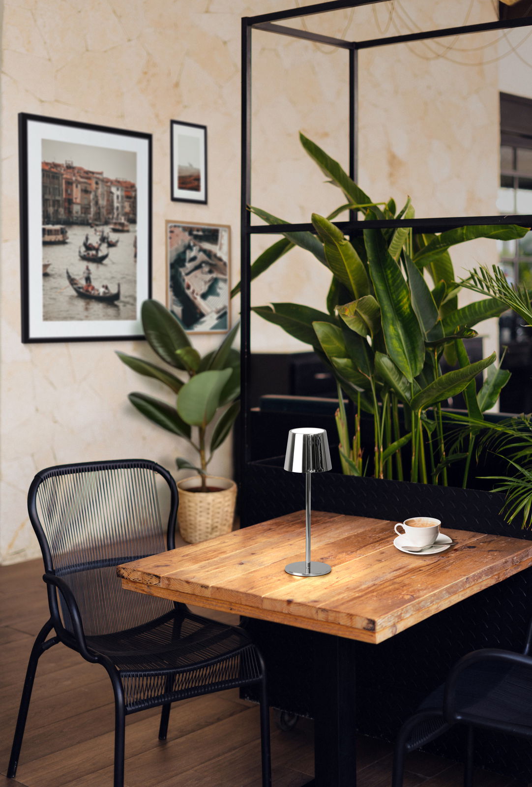 Moderne Akku-Tischleuchte ROXI auf einem Holztisch in einem stilvollen Café mit Pflanzen im Hintergrund, seitliche Perspektive.