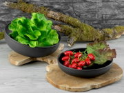 Zwei matte schwarze Servierschalen auf Holzplatten, gefüllt mit grünem Blattsalat sowie Cherrytomaten und rotem Salat, fotografiert in leichter Aufsicht vor grauer Steinwand.