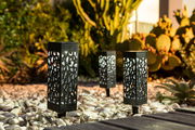 Drei dekorative Solarleuchten mit filigranem Muster, aufgestellt in einem Garten mit Kieselsteinen und Kakteen, aus seitlicher Perspektive fotografiert.