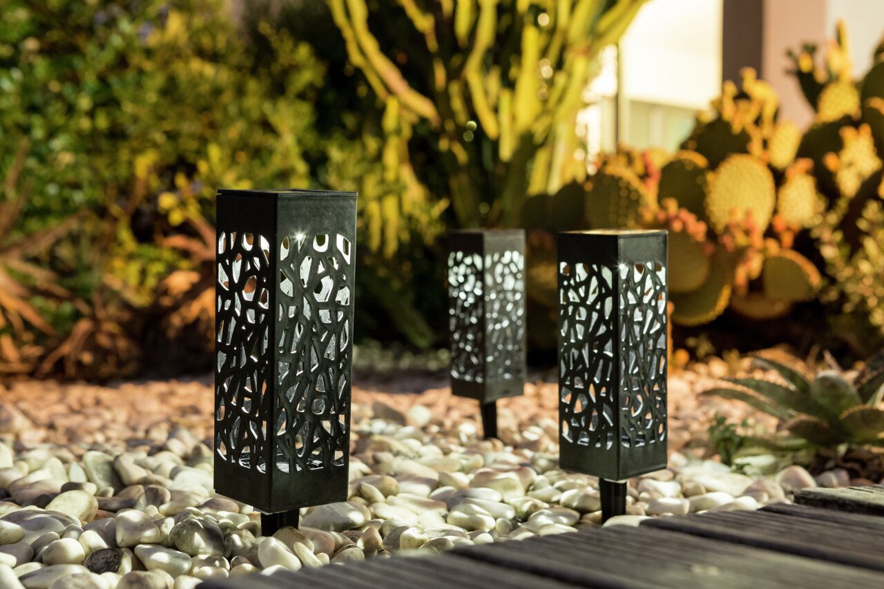 Ambia Garden Solarleuchten Set 3-tlg. Drei dekorative Solarleuchten mit filigranem Muster, aufgestellt in einem Garten mit Kieselsteinen und Kakteen, aus seitlicher Perspektive fotografiert.