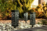 Drei dekorative Solarleuchten mit filigranem Muster, aufgestellt in einem Garten mit Kieselsteinen und Kakteen, aus seitlicher Perspektive fotografiert.