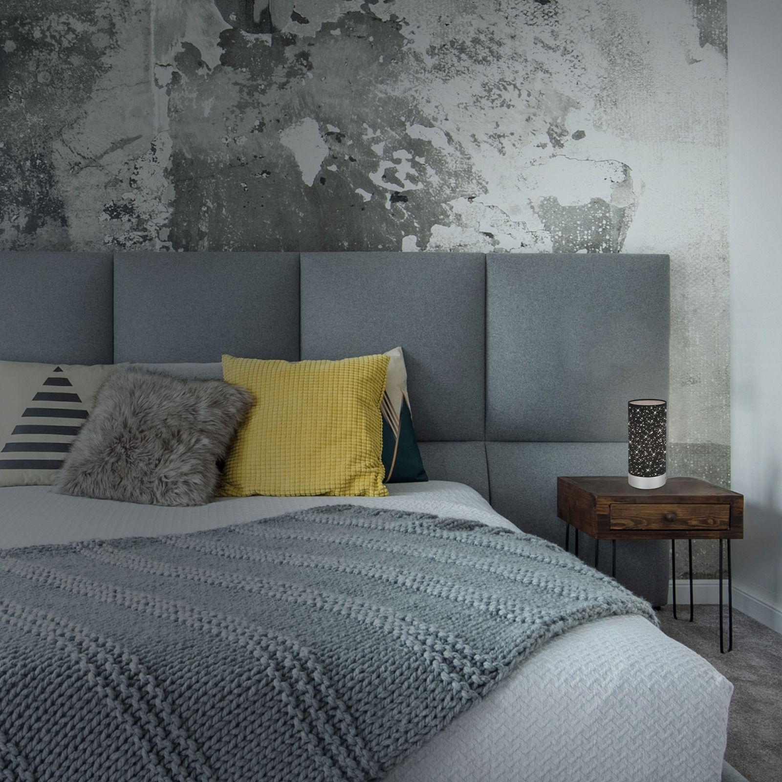 Tischleuchte auf einem Nachttisch neben einem Bett mit grauem Kopfteil, dekoriert mit Kissen und einer gestrickten Decke, aus seitlicher Perspektive.