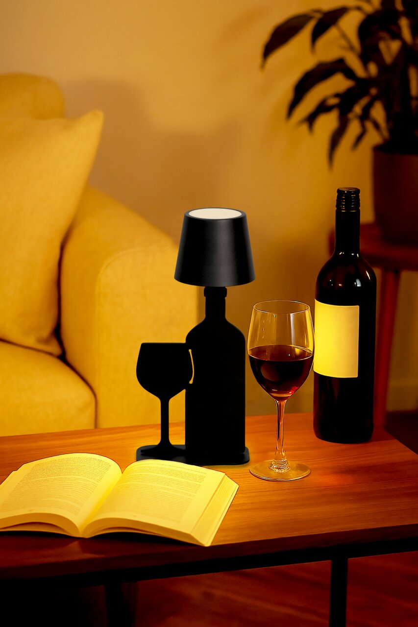Schwarze Akku-Tischleuchte in Form einer Weinflasche und eines Weinglases, auf einem Tisch neben einem Buch und einem Weinglas, aus seitlicher Perspektive.