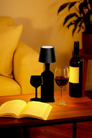 Schwarze Akku-Tischleuchte in Form einer Weinflasche und eines Weinglases, auf einem Tisch neben einem Buch und einem Weinglas, aus seitlicher Perspektive.