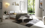 Modernes Schlafzimmer mit einem weißen Bett mit Holz- und Metallkopfteil, seitlicher Blick. Das Zimmer ist hell und minimalistisch eingerichtet mit großen Fenstern, einem Nachttisch und stilvollen Dekorationen.