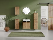 Moderner Waschbeckenunterschrank aus Holz mit vertikalen Lamellen, frontal vor einer grünen Wand montiert, umgeben von passenden Schränken und einem runden Spiegel.