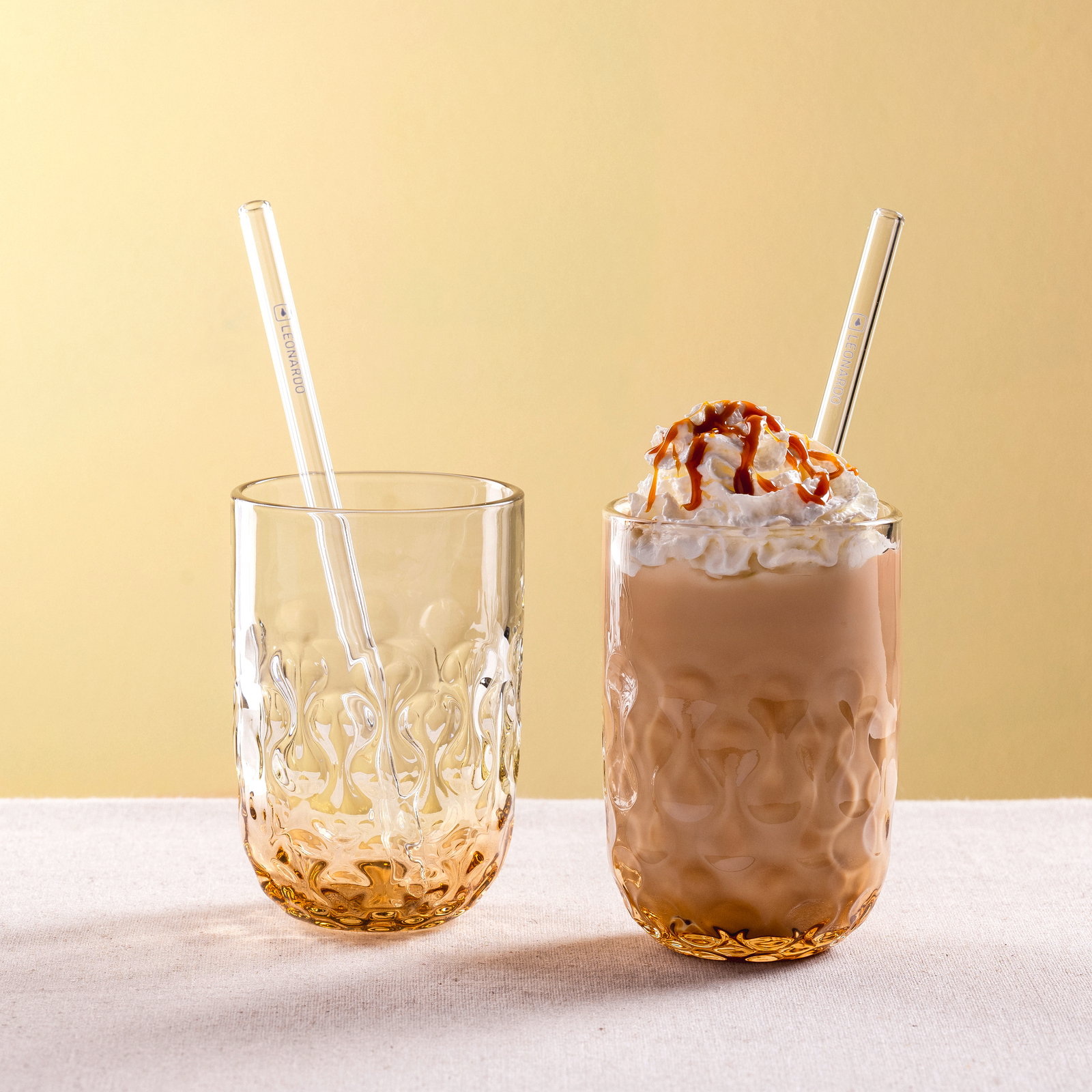 Zwei verzierte Gläser mit Strohhalmen, eines leer und eines gefüllt mit Caramel Macchiato, von vorne betrachtet.