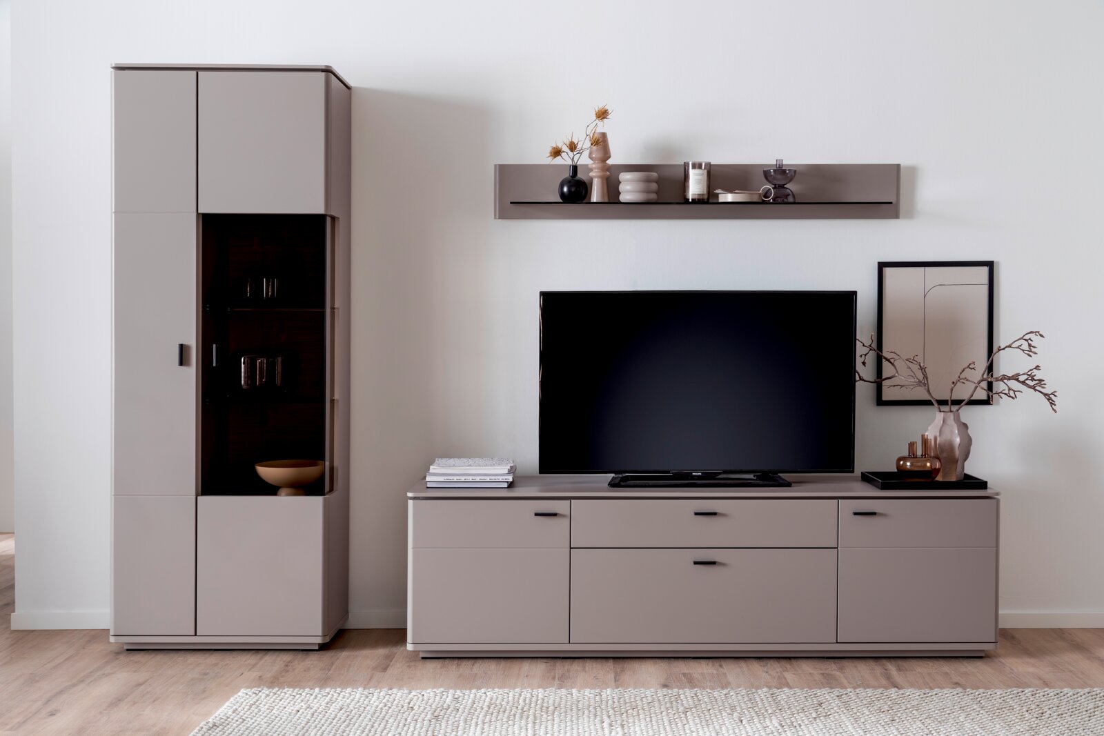 Zurbrüggen Combinatievitrine TORRENT Moderne woonkameropstelling met een taupekleurige tv-wand: links een hoge vitrinekast met glazen vakken, rechts een laag tv-lowboard met televisie en daarboven een wandplank met decoratie, gezien van voren vanuit een licht schuin perspectief.