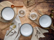 Drei hellgraue Keramikschalen mit dunklem Rand auf rustikalem Holztisch, arrangiert mit Holzbrettchen, Leinenservietten und goldfarbenem Besteck, Draufsicht (Top-Down)