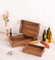 Drei hölzerne Tabletts in verschiedenen Größen, auf einem Tisch arrangiert, mit einer dekorativen Vase und Gewürzflaschen im Hintergrund. Perspektive von oben.