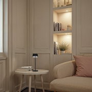 Schwarze, moderne Tischleuchte mit warmweißem Licht auf rundem Beistelltisch neben beigem Sofa, seitliche Perspektive im Wohnzimmer